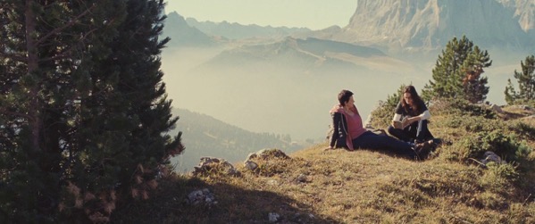 two women rest at the edge of a cliff, with Alps in the background; still from "Clouds of Sils Maria"
