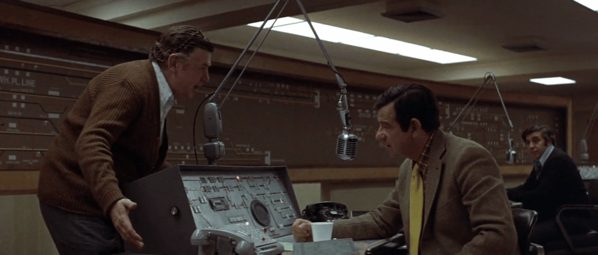 two men talk in a dispatch control room full of computers and microphones; still from "The Taking of Pelham One Two Three"